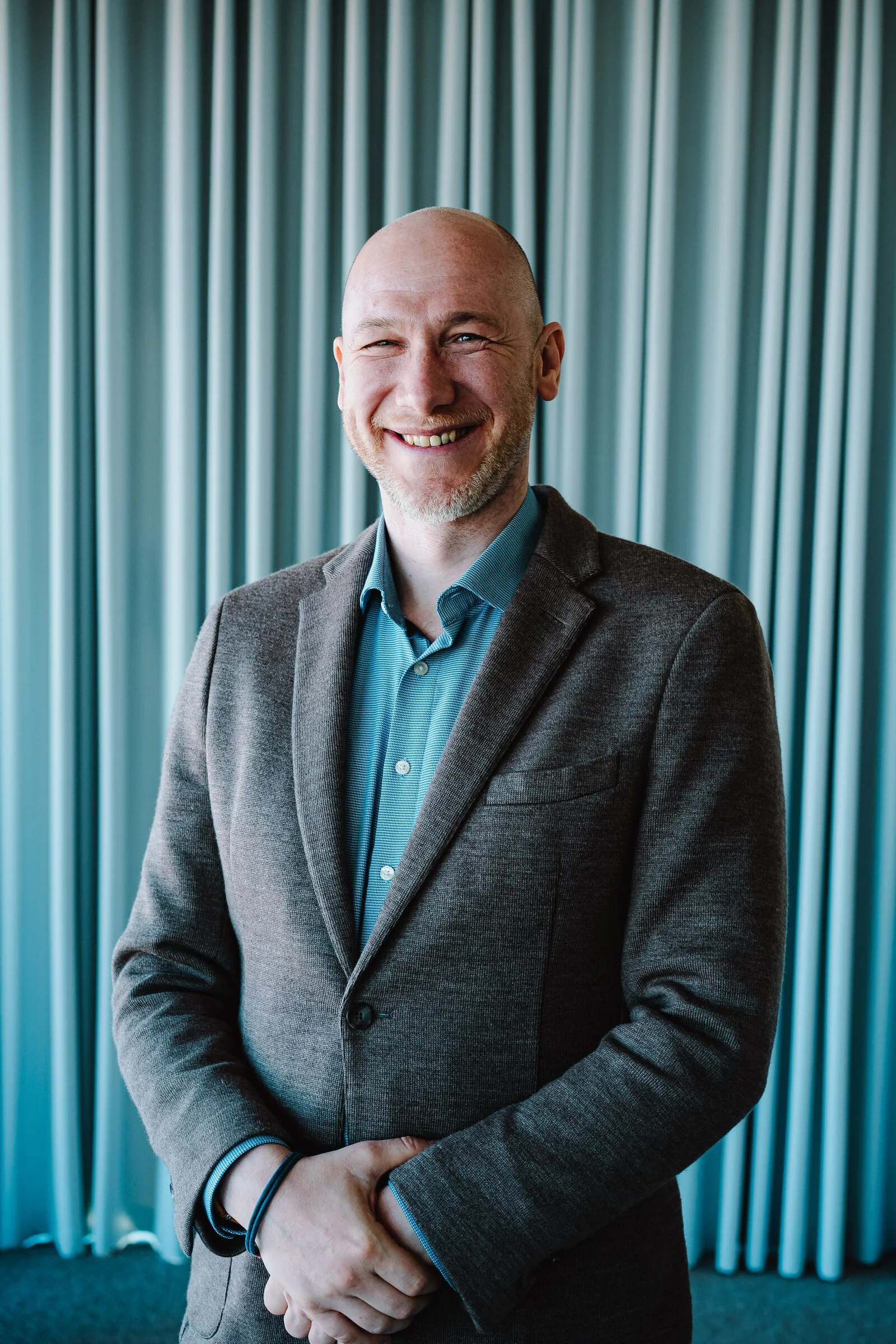 Marian Kamenistak in a grey blazer against a teal curtain, warm smile.