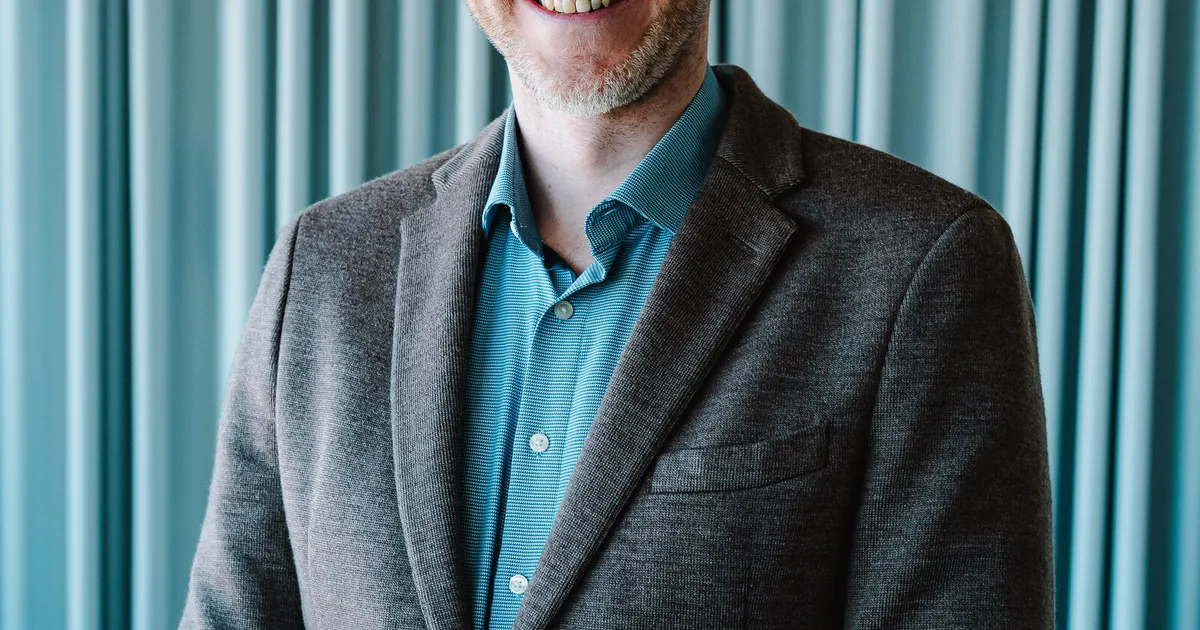Marian Kamenistak in a grey blazer against a teal curtain, warm smile.