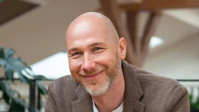 Marian Kamenistak nakloněný dopředu v hnědém saku, usmívá se.