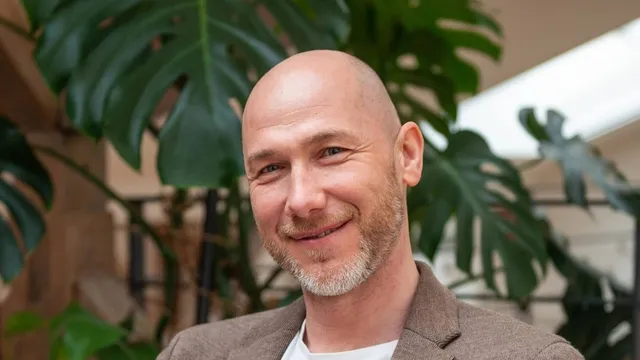 Marian Kamenistak smiling at the camera in a brown blazer, large monstera leaves behind him.