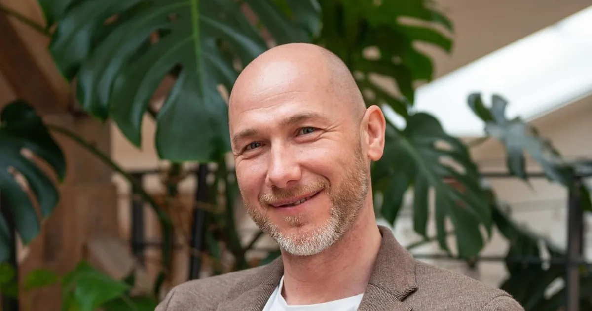 Marian Kamenistak smiling at the camera in a brown blazer, large monstera leaves behind him.