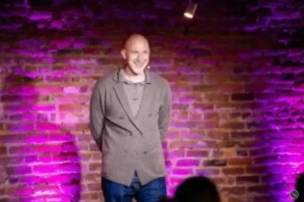 Marian Kamenistak on a brick-walled venue stage lit in purple, smiling at the audience.