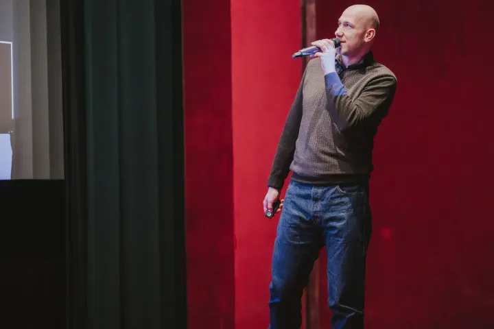 Marian Kamenistak speaking with a microphone against a red velvet curtain backdrop.