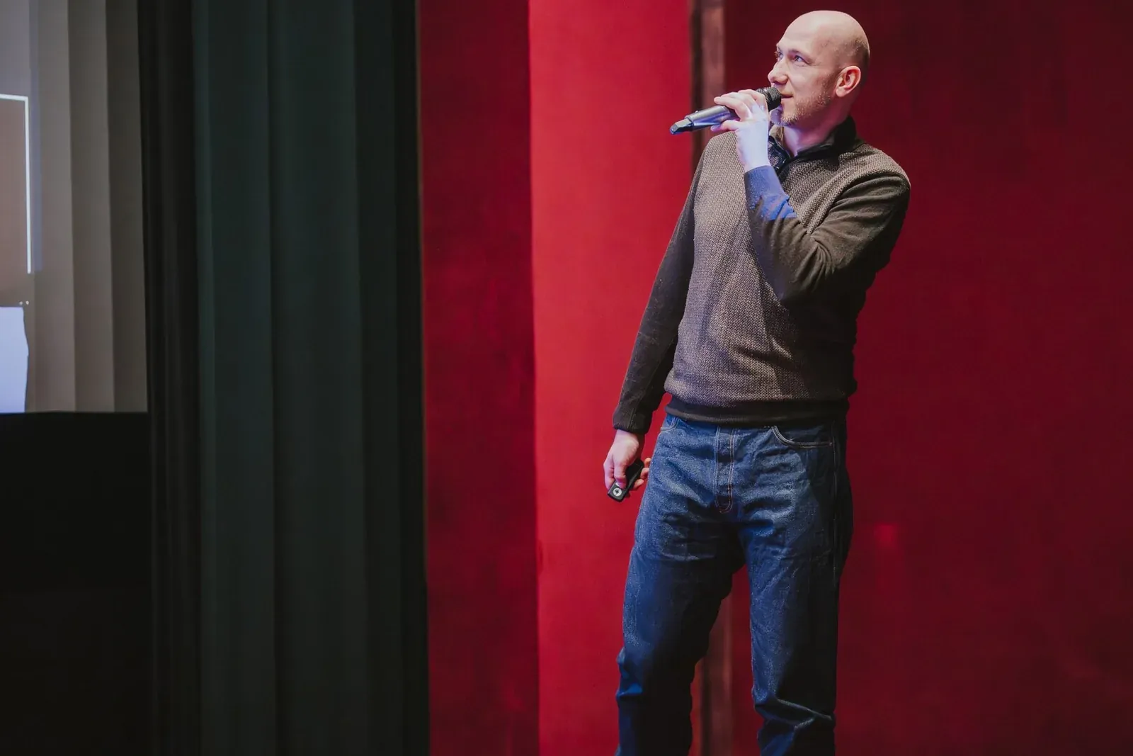 Marian Kamenistak speaking with a microphone against a red velvet curtain backdrop.