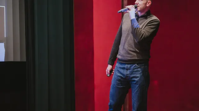Marian Kamenistak speaking with a microphone against a red velvet curtain backdrop.