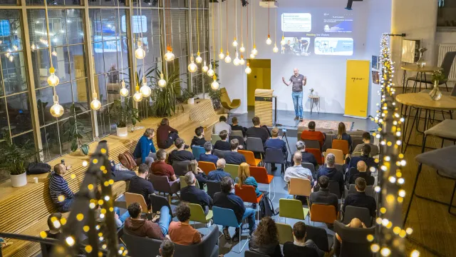 Wide shot of Marian Kamenistak speaking to a seated audience in a light-strung venue.