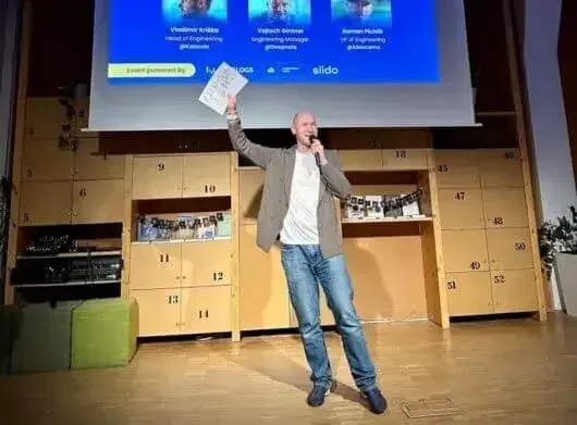 Marian Kamenistak on stage holding a printed panel with speaker photos projected behind him.