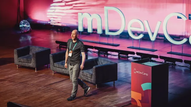Marian Kamenistak walking the mDevCamp stage in front of a large pink-lit conference backdrop.