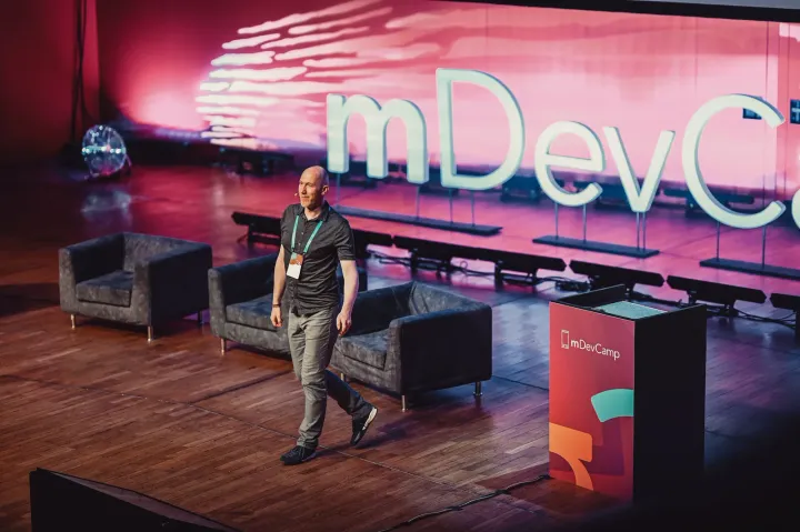 Marian Kamenistak walking the mDevCamp stage in front of a large pink-lit conference backdrop.