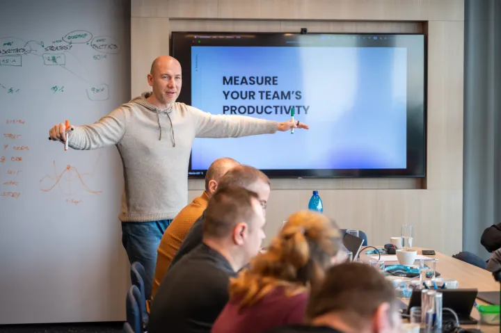 Marian Kamenistak leading a workshop in front of a screen reading "Measure your team's productivity" with strategy notes on the whiteboard.