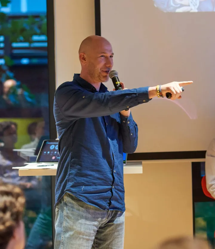 Marian Kamenistak in a denim shirt speaking into a handheld microphone and pointing at someone in the audience, a projector screen behind him.