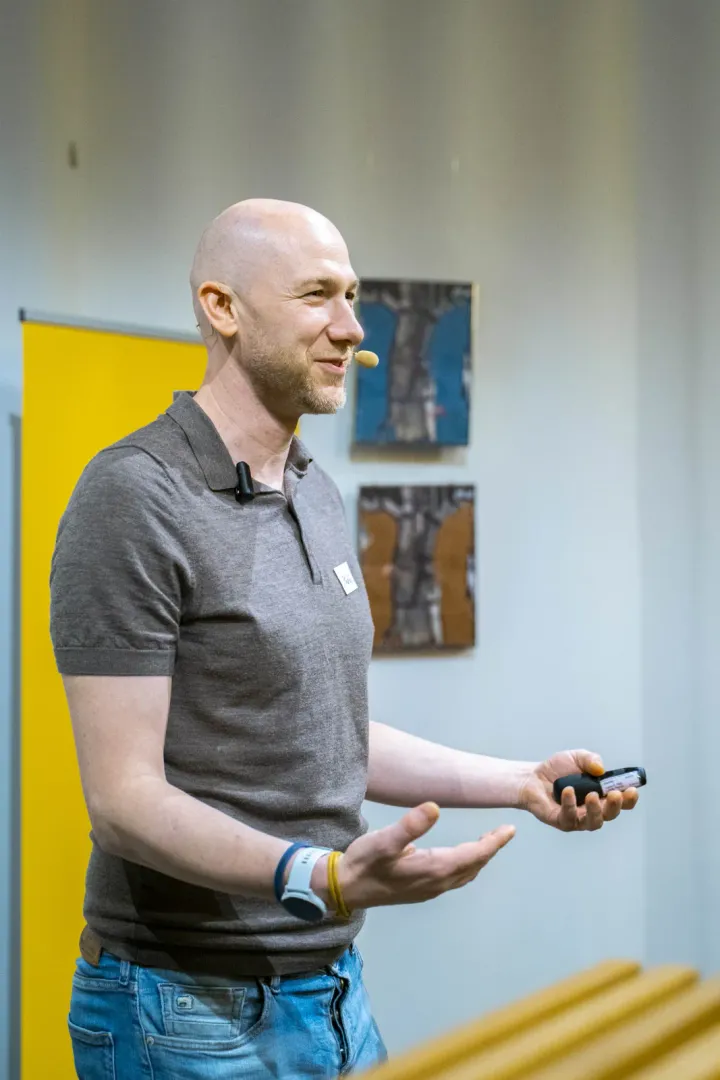 Marian Kamenistak in a grey polo shirt with a headset microphone, smiling and explaining a point with open hands, a presentation clicker in his right hand.