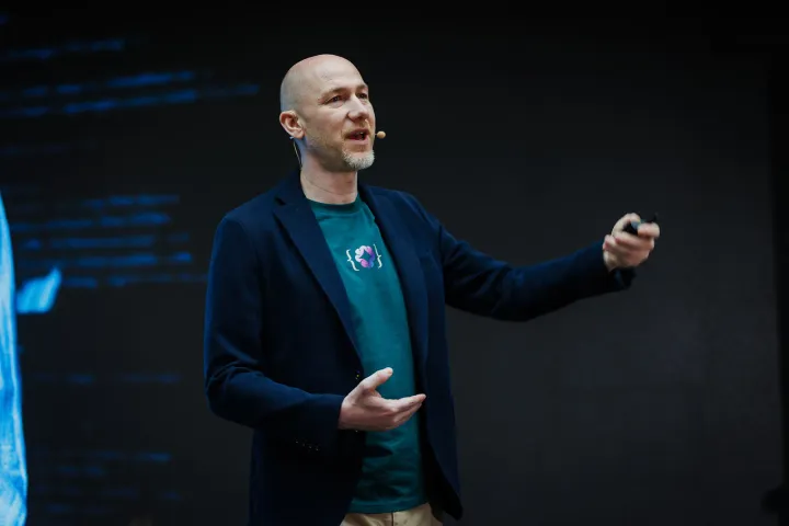 Marian Kamenistak mid-talk on a dark conference stage, lines of code projected on the screen behind him.
