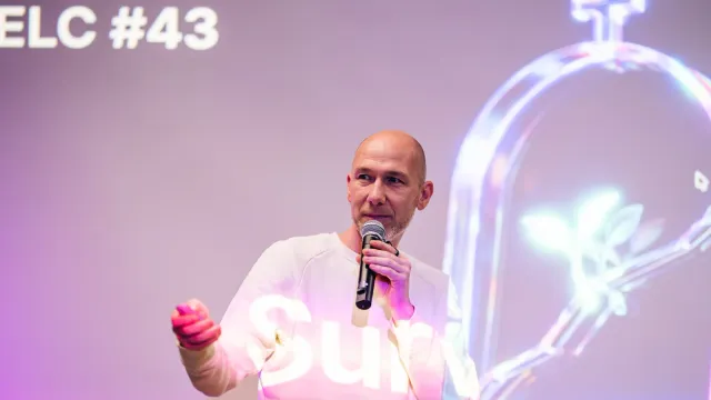 Marian Kamenistak na pódiu s mikrofonem gestikuluje k publiku pod úvodním slidem ELC #43.