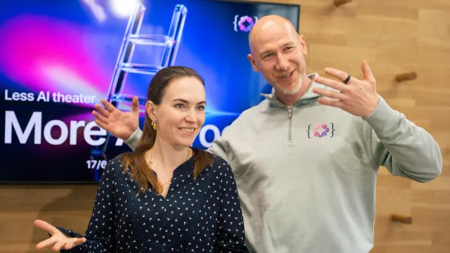 Marian Kamenistak gestikuluje vedle usměvavé hostky před obrazovkou s nápisem „Less AI theater“ na meetupu ELC.