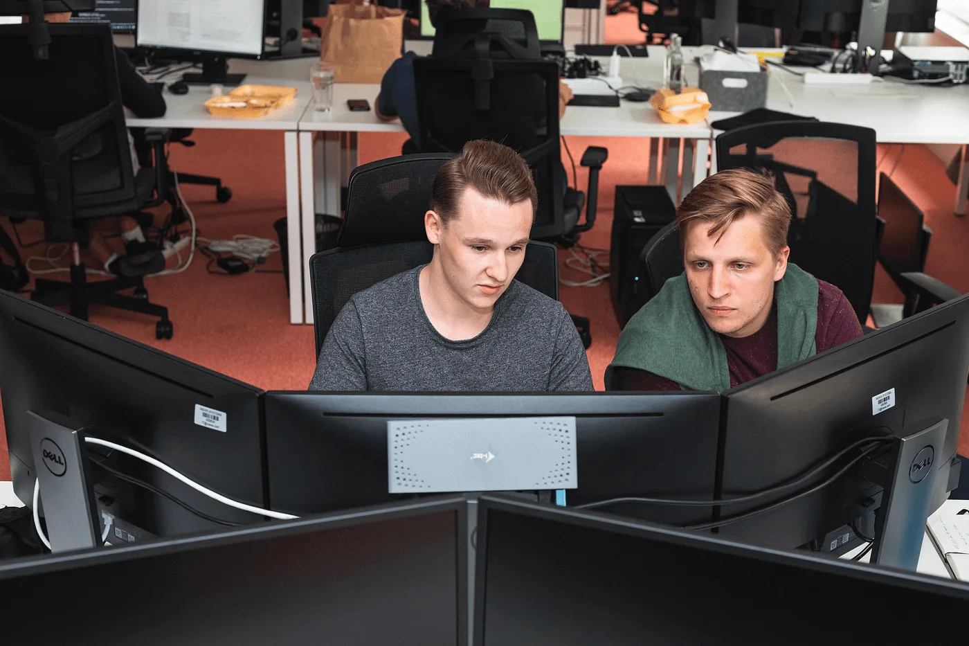 Two developers sharing one screen at a desk in an open-plan office, working through a problem