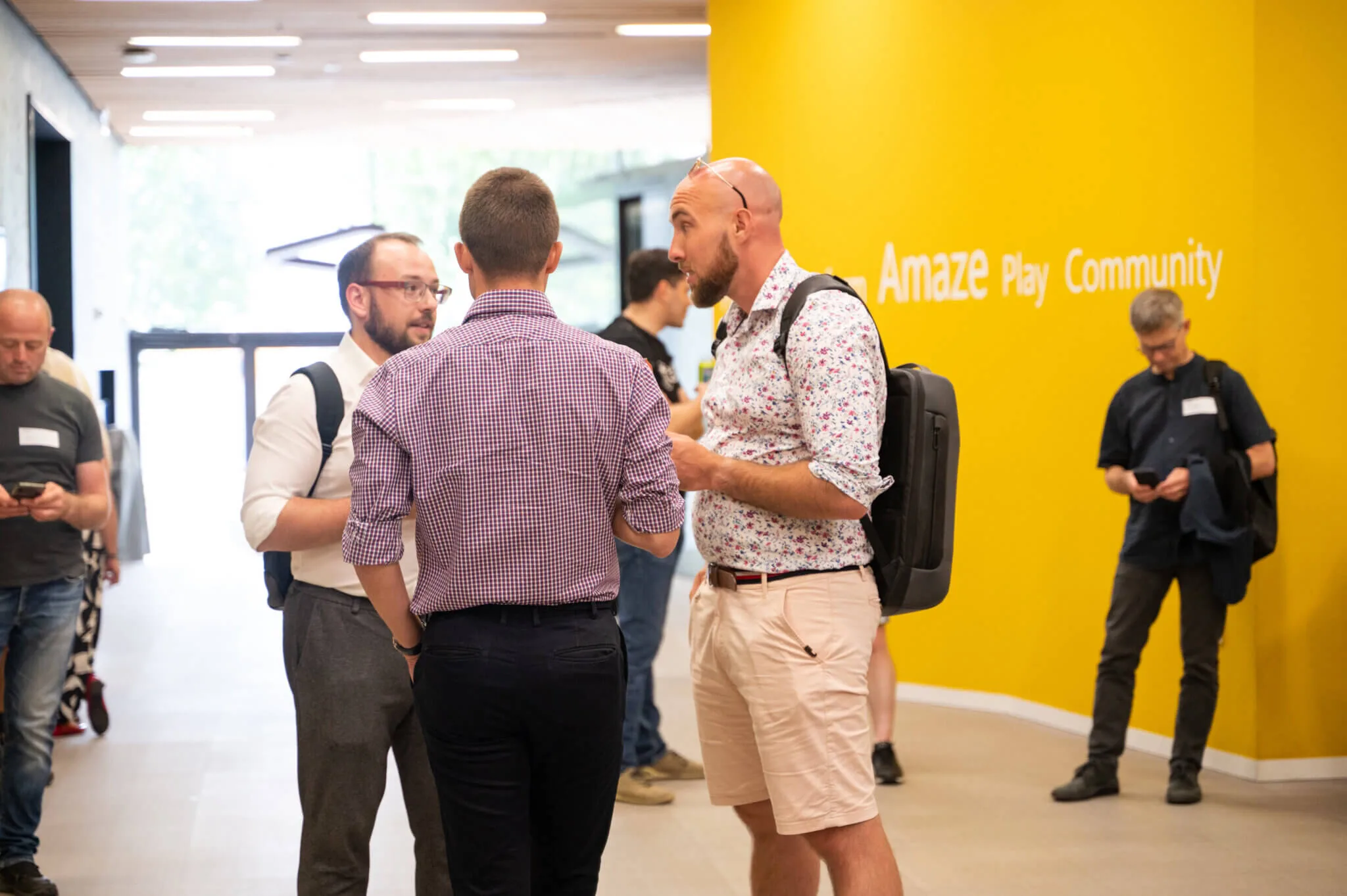Attendees with backpacks talking in a corridor by a yellow wall reading Amaze Play Community