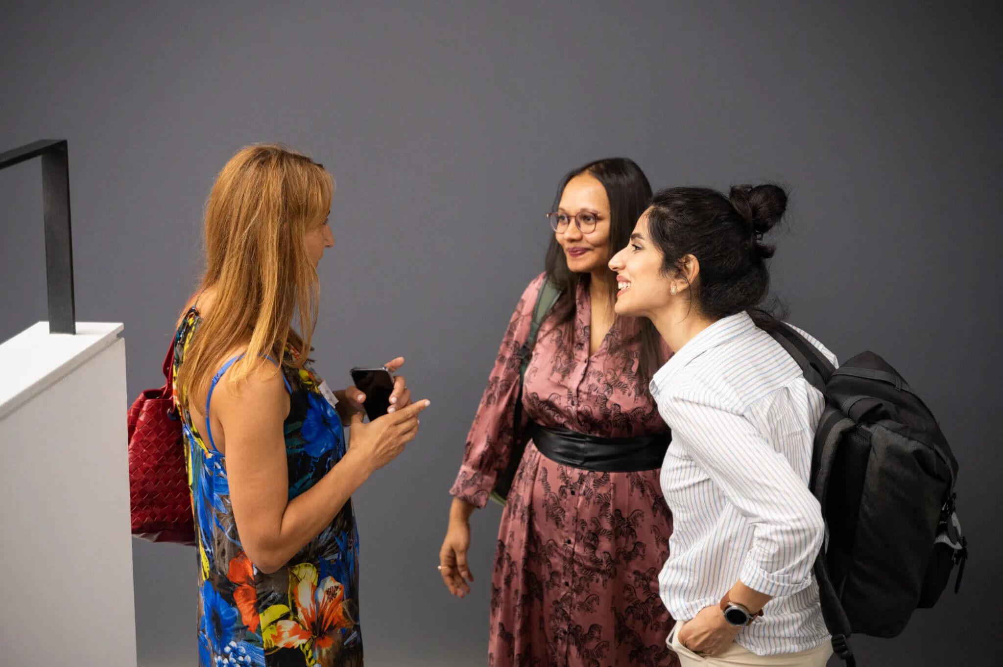 Three women in conversation at an ELC meetup, one showing something on her phone against a grey wall