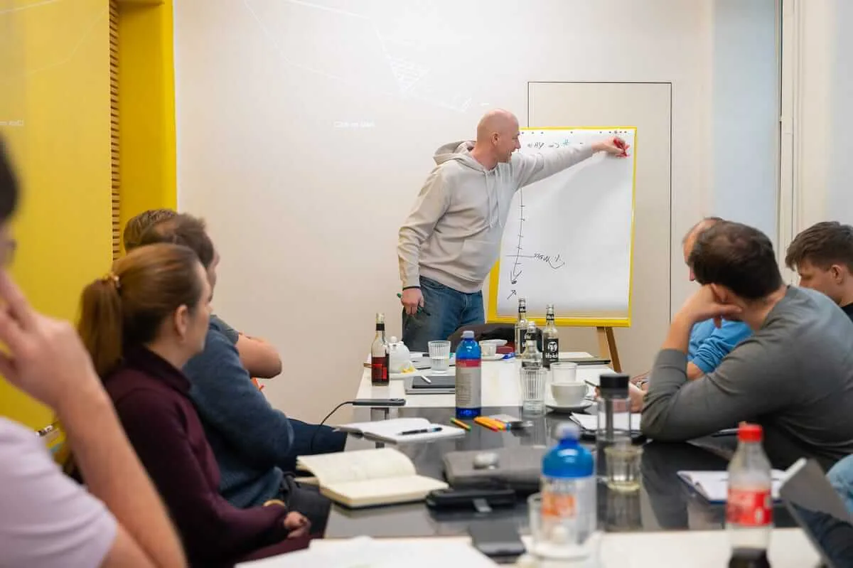A trainer sketching a chart on a flipchart while course participants follow from around a boardroom table
