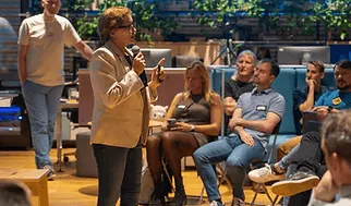A speaker in a light blazer with a handheld microphone addressing seated attendees at an ELC community meetup