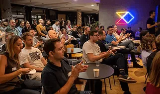 Attendees seated at small tables listening at an ELC meetup, a neon diamond sign glowing on the wall behind